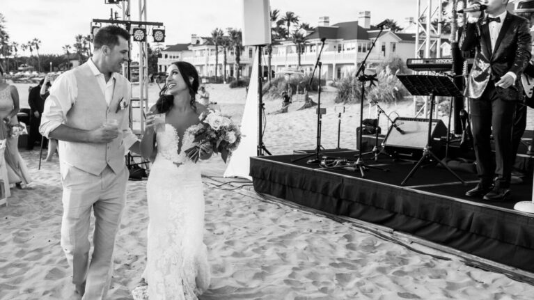 A joyful couple walks hand-in-hand along the beach, celebrating their wedding day at Hotel Del Coronado as a live wedding band performs in the background. The scene captures the vibrant atmosphere created by the 20-piece wedding band, enhancing the romantic setting. This image reflects the exceptional entertainment provided by Ben Mallare Events & Entertainment, perfect for making unforgettable memories on your special day.