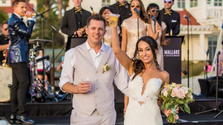 A joyful couple celebrates their wedding on the beach at Hotel Del Coronado, raising glasses in front of a vibrant 20-piece live wedding band performing on stage. The picturesque setting showcases Ben Mallare Events & Entertainment, highlighting the energetic atmosphere that live music brings to wedding celebrations. The musicians are dressed in formal attire, creating an elegant backdrop for this memorable moment.