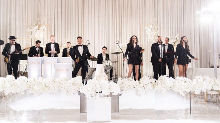 A 20-piece live wedding band from Ben Mallare Events & Entertainment performing on stage, featuring musicians and singers in elegant attire, with a backdrop of luxurious white floral arrangements. A 20-piece live wedding band from Ben Mallare Events & Entertainment performing on stage, featuring musicians and singers in elegant attire, with a backdrop of luxurious white floral arrangements.
