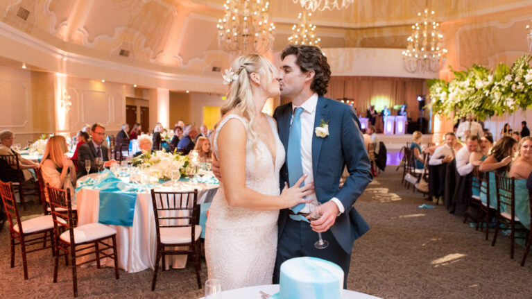 A couple shares a romantic kiss during their wedding reception in the Ballroom at Hotel Del Coronado, surrounded by elegantly arranged tables and guests enjoying the celebration. In the background, a lively 20-piece wedding band performs, enhancing the festive atmosphere of this Ben Mallare Events & Entertainment event. This image captures the joy and energy that live wedding bands bring to special occasions, making them an essential element for unforgettable weddings.