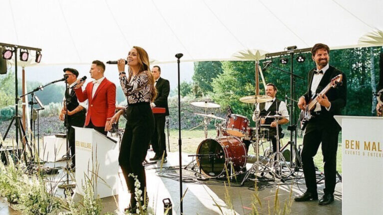 Live wedding band performing on stage, featuring multiple musicians including a singer, guitarist, drummer, and keyboardist, under a white canopy. Ben Mallare Events & Entertainment 20-piece wedding band providing live entertainment at an outdoor wedding.