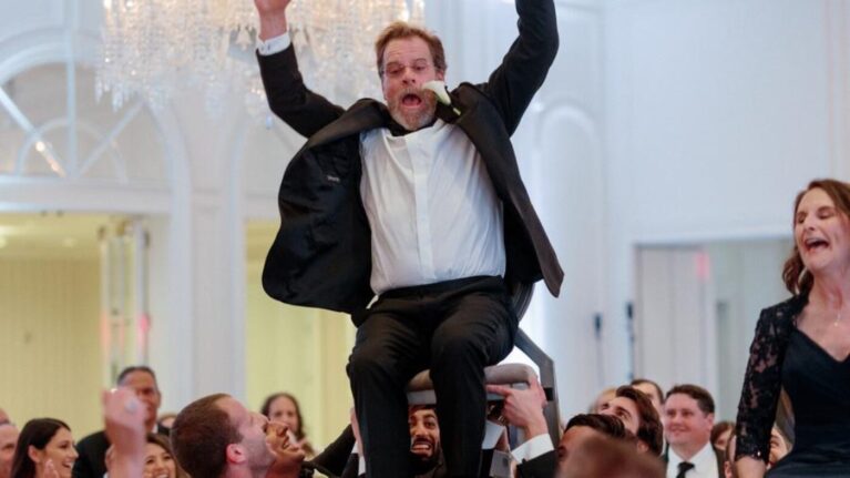 A lively wedding celebration at Rosewood Miramar Beach featuring guests joyfully lifting a groom on a chair, surrounded by friends and family. The elegant venue showcases a beautiful chandelier, enhancing the festive atmosphere. This moment highlights the excitement and energy that live wedding bands, like those from Ben Mallare Events & Entertainment, bring to special occasions, creating unforgettable memories on the dance floor.