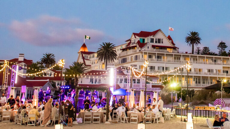 A vibrant beach wedding at Hotel Del Coronado featuring a lively 20-piece wedding band performing on stage, surrounded by elegantly dressed guests enjoying a beautiful evening. The picturesque backdrop of the coastal venue, complemented by string lights and palm trees, enhances the festive atmosphere. This image showcases the perfect setting for memorable celebrations, highlighting the quality of live wedding bands provided by Ben Mallare Events & Entertainment.