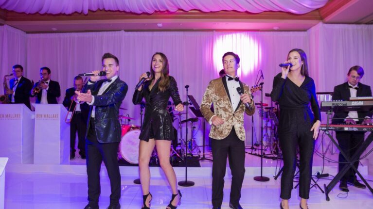 A 20-piece live wedding band from Ben Mallare Events & Entertainment performing on stage, featuring four vocalists dressed elegantly, with musicians playing instruments in the background, under vibrant event lighting.