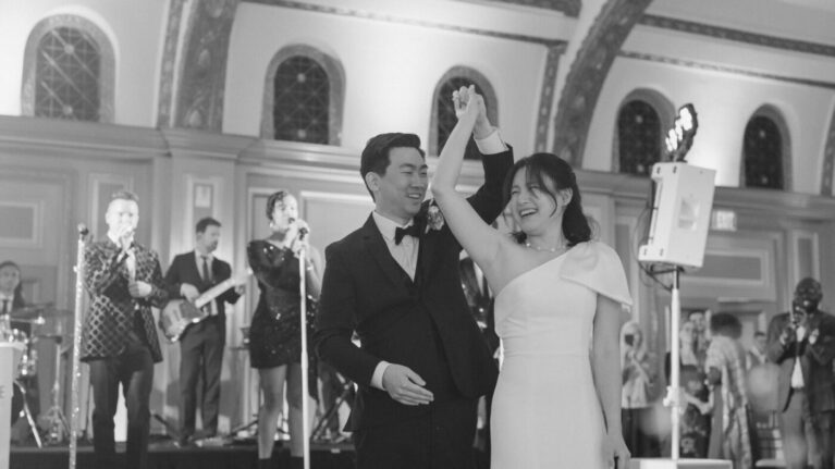 A joyful couple dances together at their wedding reception at the Langham Huntington, Pasadena, surrounded by a lively 20-piece wedding band performing on stage. The elegant venue features a stunning chandelier, creating a romantic atmosphere. This moment captures the essence of live wedding bands, enhancing the celebration with unforgettable music and energy, perfect for any special event. Ben Mallare Events & Entertainment specializes in creating memorable experiences with talented musicians.