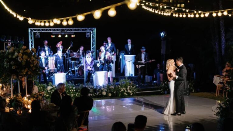 A 20-piece live wedding band from Ben Mallare Events & Entertainment performs on stage under string lights, while a couple shares their first dance at an elegant outdoor wedding reception.