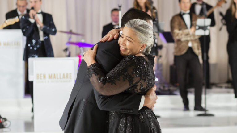 A heartfelt moment captured during a wedding reception featuring live music from a 20-piece wedding band by Ben Mallare Events & Entertainment. The image shows a joyful embrace between a couple, with musicians performing in the background, highlighting the vibrant atmosphere created by live wedding bands. Perfect for couples seeking an unforgettable musical experience for their special day.