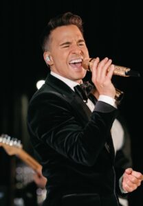 A charismatic male vocalist performs on stage, showcasing his talent as part of a dynamic 20-piece wedding band. The energy of the live wedding music creates an unforgettable atmosphere, ideal for making special memories at events like those organized by Ben Mallare Events & Entertainment.