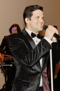 A charismatic lead singer performs on stage with a 20-piece wedding band, captivating guests at a wedding reception. The musicians, dressed in stylish attire, create an energetic atmosphere, showcasing the vibrant entertainment options available for special events. Ben Mallare Events & Entertainment specializes in live wedding bands, ensuring unforgettable musical experiences tailored to every celebration.