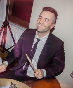 A smiling drummer performing with a vibrant 20-piece wedding band at an elegant wedding reception, showcasing the energetic atmosphere and live entertainment from Ben Mallare Events & Entertainment. The image captures the dynamic interaction between musicians, highlighting the memorable experience of live wedding music that creates unforgettable moments for couples and their guests.