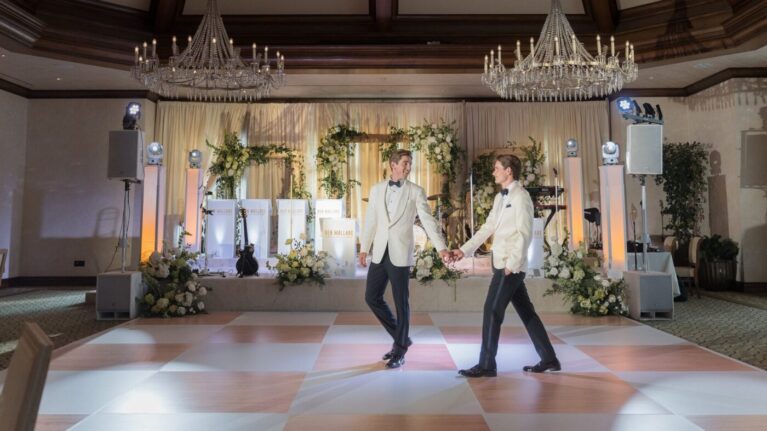 Two elegantly dressed grooms dance hand-in-hand on a beautifully lit dance floor at their wedding reception, surrounded by floral arrangements and a stage set for a live wedding band performance. The backdrop features the professional setup of Ben Mallare Events & Entertainment, showcasing a polished 20-piece wedding band ready to create an unforgettable atmosphere for the couple and their guests. This image captures the joy and elegance of a celebration enhanced by live music, perfect for couples looking to elevate their wedding experience.