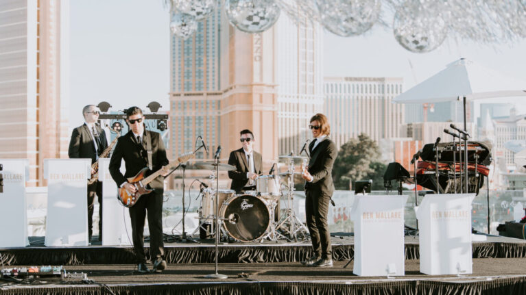 Live wedding band from Ben Mallare Events & Entertainment performing on stage with a 20-piece ensemble, featuring musicians playing guitars, drums, and keyboards, set against a scenic city backdrop.