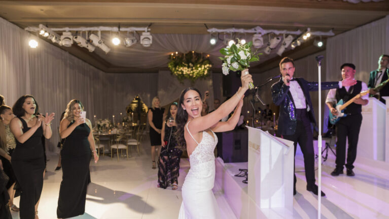 A lively moment captured at a wedding reception at the Resort at Pelican Hill featuring a joyful bride celebrating with a bouquet in hand, surrounded by guests enjoying the performance of a talented 20-piece wedding band. The scene showcases the vibrant atmosphere created by live wedding bands, emphasizing the energy and excitement they bring to special events. Ben Mallare Events & Entertainment specializes in providing unforgettable musical experiences for weddings, ensuring memorable celebrations filled with dancing and fun.