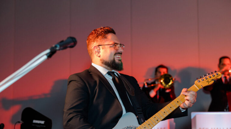 Live wedding band guitarist performing on stage with a trumpet player in the background, part of a 20-piece wedding band from Ben Mallare Events & Entertainment.