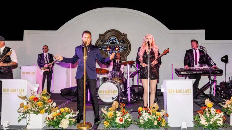 Live wedding band from Ben Mallare Events & Entertainment performing on stage with a 20-piece ensemble, featuring male and female lead singers, drummer, and guitarist, surrounded by floral decorations.