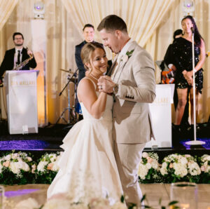 A couple shares a romantic dance during their wedding reception, surrounded by a vibrant 20-piece wedding band from Ben Mallare Events & Entertainment. The band features talented musicians performing live, creating an unforgettable atmosphere for the celebration. Elegant floral arrangements and soft lighting enhance the festive ambiance, making it a perfect setting for a memorable wedding experience.