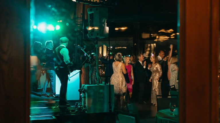 A lively scene captured through a wooden door at the Crown Room at Hotel Del Coronado showcasing a 20-piece wedding band performing at a reception, with guests dancing and enjoying the vibrant atmosphere. The musicians are set against a backdrop of colorful lights, highlighting the energy of live wedding entertainment. Ben Mallare Events & Entertainment provides unforgettable musical experiences, ensuring that your special day is filled with joy and celebration.