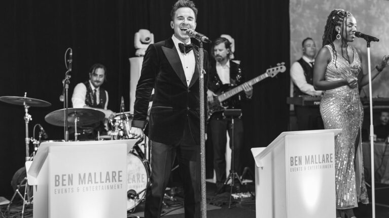 A lively performance by a 20-piece wedding band at Hotel Del Coronado, featuring talented musicians including a male vocalist in a tuxedo and a female singer in a sparkling gown. The band, associated with Ben Mallare Events & Entertainment, showcases a vibrant atmosphere perfect for weddings, ensuring an unforgettable entertainment experience for guests.