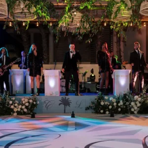 A vibrant live wedding band performance by Ben Mallare Events & Entertainment featuring multiple musicians on stage, including vocalists and instrumentalists, creating a memorable atmosphere for wedding celebrations. The 20-piece ensemble showcases a captivating mix of music, surrounded by elegant floral arrangements and dynamic stage lighting, enhancing the festive ambiance for couples and their guests.